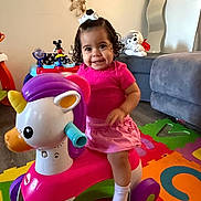 Antonielys is registered to the contest to win money with this photo: toddler, child, unicorn_toy, play_mat, pink_clothing, white_socks, curly_hair, bow, smiling, indoor, living_room, plush_toy, sofa, mirror, wooden_floor, colorful, happy, cute, toy, playtime