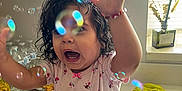Antonielys is registered to the contest to win money with this photo: toddler, child, bubbles, curly_hair, flower_headband, pink_shirt, red_pants, kitchen, countertop, window, daylight, happy, playful, indoor, water_bottles, green_cloth, reaching_hands, cute, smiling, person