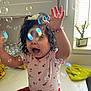 toddler, child, bubbles, curly_hair, flower_headband, pink_shirt, red_pants, kitchen, countertop, window, daylight, happy, playful, indoor, water_bottles, green_cloth, reaching_hands, cute, smiling, person