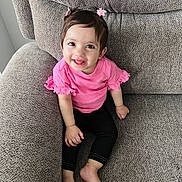 Sanvi is registered to the contest to win money with this photo: barefoot, black_pants, bright, casual, child, couch, cozy, cute, furniture, hair_bows, happy, home, indoor, person, pink_top, portrait, sitting, smiling, toddler, young_child
