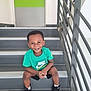 child, boy, smiling, stairs, green_tshirt, nike, black_shorts, black_shoes, white_socks, indoors, door, railing, happy, portrait, casual_clothing, young_child, sitting, concrete_stairs, modern, playful