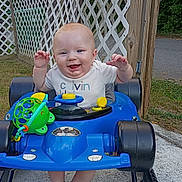 Scott is registered to the contest to win money with this photo: baby, smiling, toy_car, blue, outdoor, fence, wooden, grass, greenery, happy, child, clothing, socks, playing, cute, person, walker, toys, face, baby_girl_or_boy
