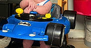 Scott is registered to the contest to win money with this photo: baby, walker, blue, batman, toy, infant, standing, arms_folded, indoor, carpet, stool, storage_bin, plastic, footwear, person, child, face, head, floor, furniture