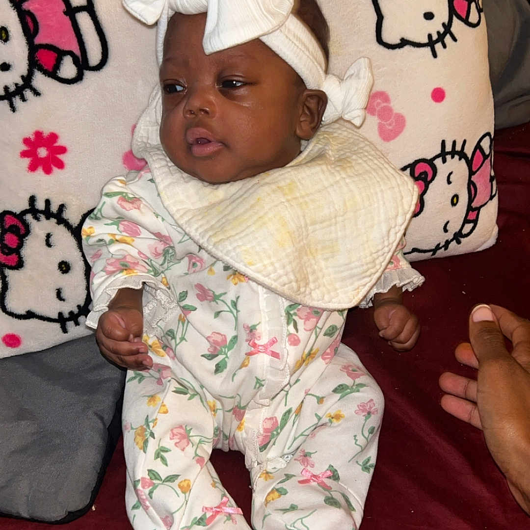 Miracle Bell joined the competition — help win amazing prizes! baby, bib, child, cloth, cute, floral_pajamas, hand, headband, hello_kitty, indoors, infant, maroon_bedspread, pattern, person, pillow, pink, portrait, sitting, soft, white