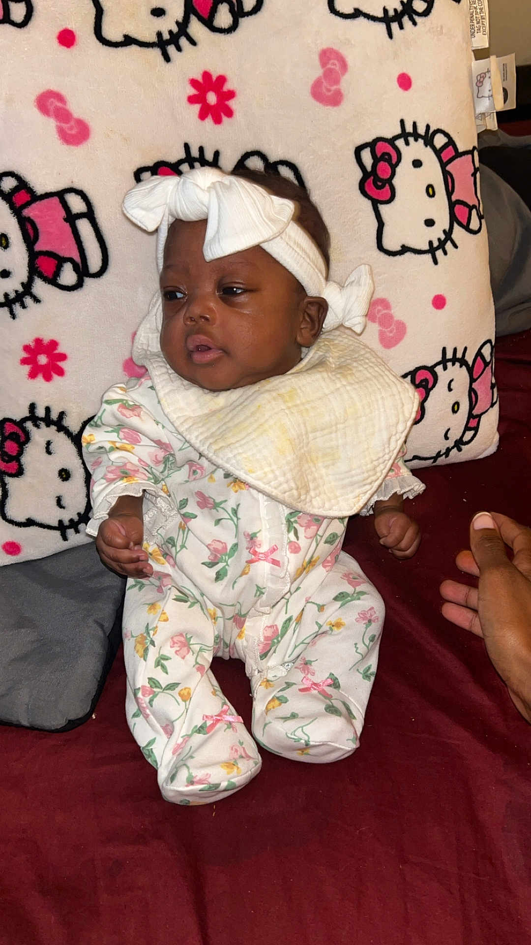 Miracle Bell joined the competition — help win amazing prizes! baby, infant, headband, bib, floral_pajamas, hello_kitty, pillow, hand, maroon_bedspread, cute, indoors, child, portrait, sitting, person, cloth, pattern, pink, white, soft
