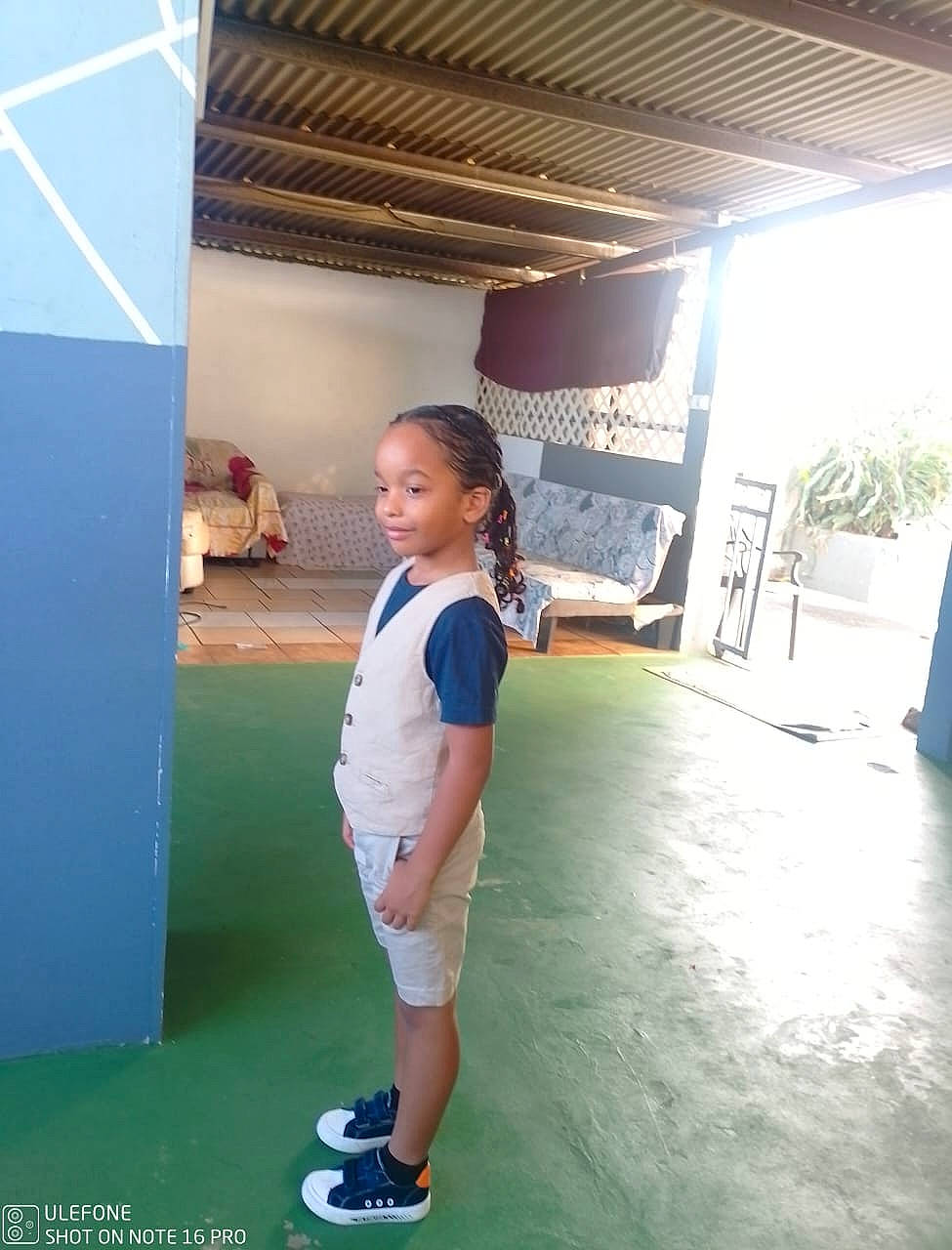Emmanuel a rejoint le concours — aidez-le/la à gagner de superbes lots ! boy, braided_hair, casual_clothing, ceiling, child, furniture, green_floor, home, indoor, natural_light, person, shorts, side_view, smile, sneakers, sofa, standing, vest, wall, young