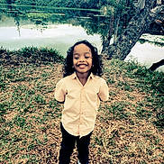 Emmanuel a rejoint le concours — aidez-le/la à gagner de superbes lots ! casual_clothing, child, curly_hair, daylight, grass, happy, lake, landscape, nature, outdoor, pants, person, reflection, scenic, shirt, smiling, sneakers, standing, tree, water