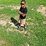 boy, child, grass, outdoor, sunglasses, black_shirt, shorts, crocs, shadow, sunlight, casual, standing, greenery, tree, bush, necklace, confident, summer, person, nature