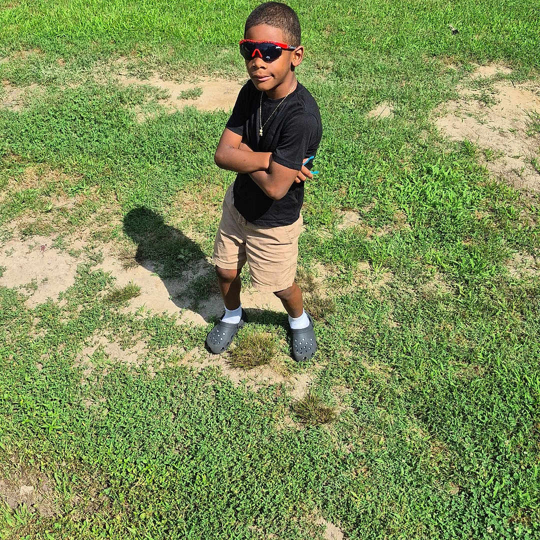 Ziyan joined the competition — help win amazing prizes! boy, child, grass, outdoor, sunglasses, black_shirt, shorts, crocs, shadow, sunlight, casual, standing, greenery, tree, bush, necklace, confident, summer, person, nature