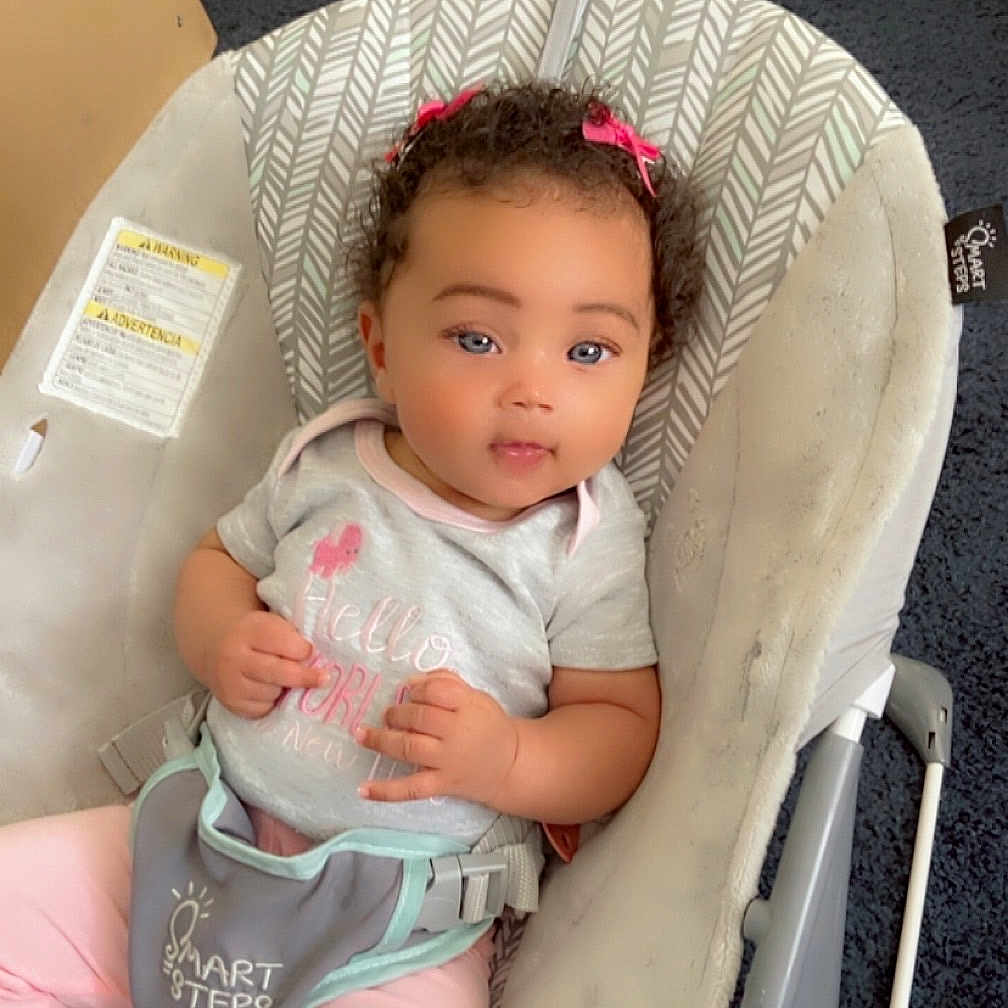 Jenesis is registered to the contest to win money with this photo: baby, child, baby_rocker, curly_hair, pink_bows, grey_shirt, pink_pants, carpet, indoor, infant, seated, cute, looking_at_camera, soft_toys, comfortable, childcare, baby_gear, face, person, young_child