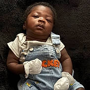 Sai is registered to the contest to win money with this photo: baby, infant, denim_overalls, mittens, black_blanket, cartoon_characters, cute, portrait, child, clothing, soft_texture, indoor, laying_down, serious_expression, person, young_child, casual_wear, closeup, newborn, small_hands