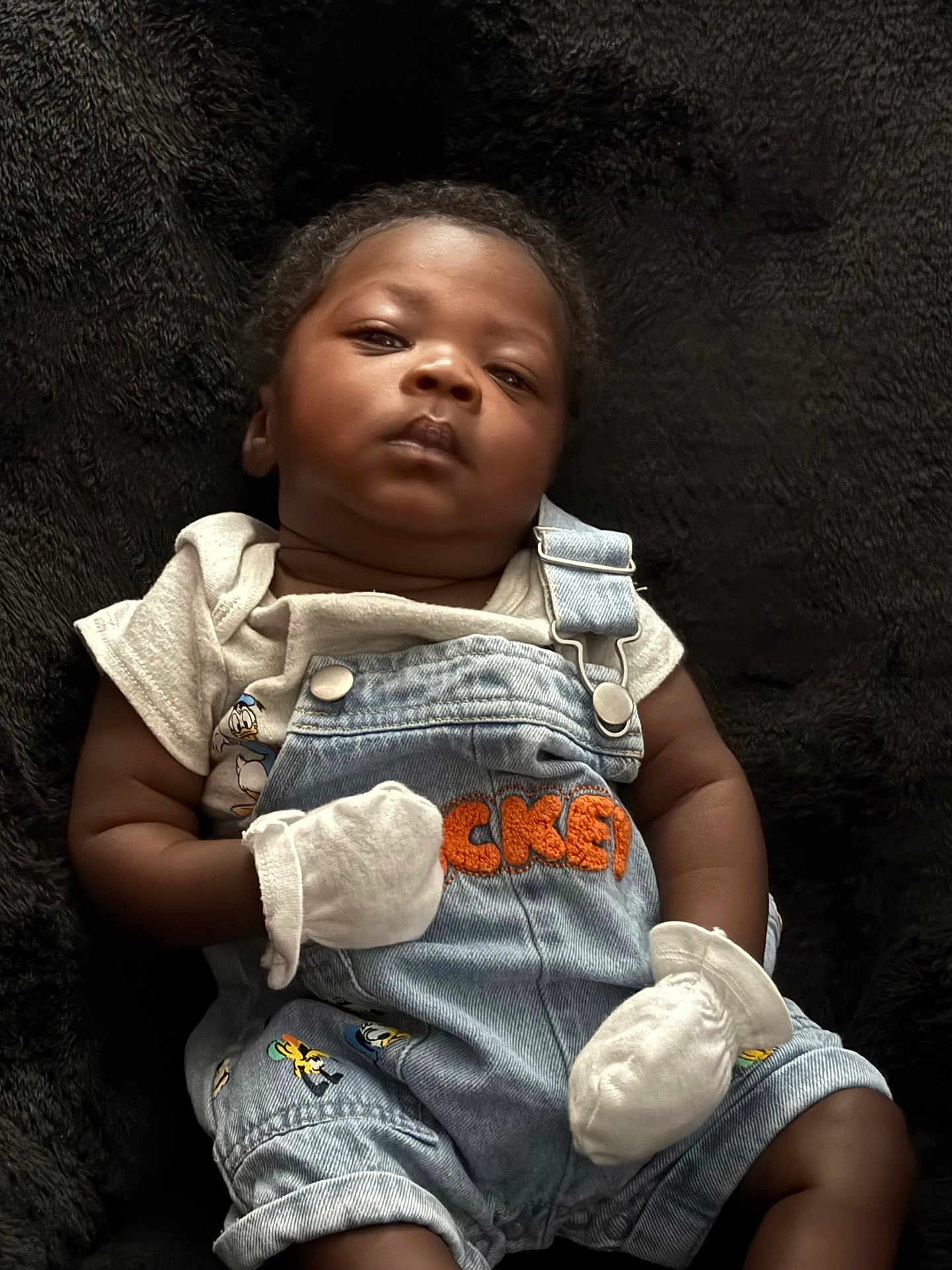 Sai is registered to the contest to win money with this photo: baby, infant, denim_overalls, mittens, black_blanket, cartoon_characters, cute, portrait, child, clothing, soft_texture, indoor, laying_down, serious_expression, person, young_child, casual_wear, closeup, newborn, small_hands