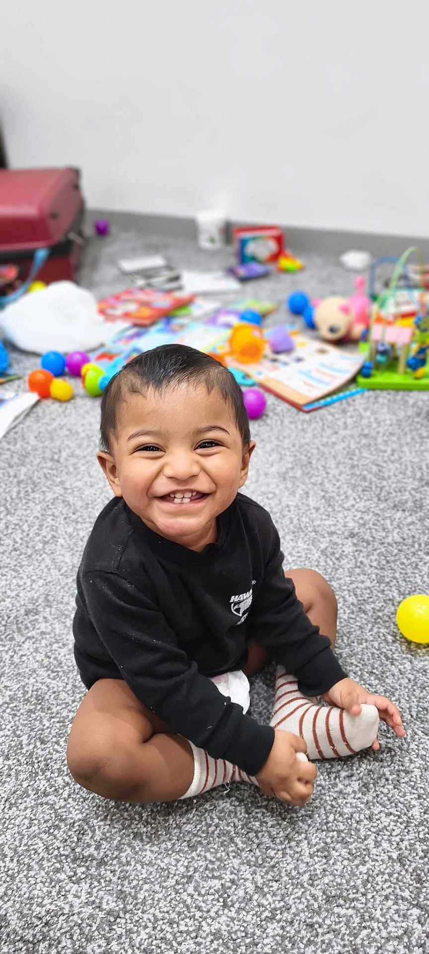 Anantajith joined the competition — help win amazing prizes! toddler, child, smiling, happy, indoor, carpet, toy, colorful, book, sitting, striped_socks, black_sweater, person, playful, cute, young_child, face, hands, feet, room