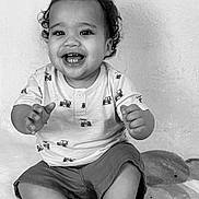 Jairo is registered to the contest to win money with this photo: baby, toddler, smiling, curly_hair, sitting, blanket, pattern, shorts, shirt, hand, feet, face, child, indoors, portrait, black_and_white, happy, cute, clothing, expression