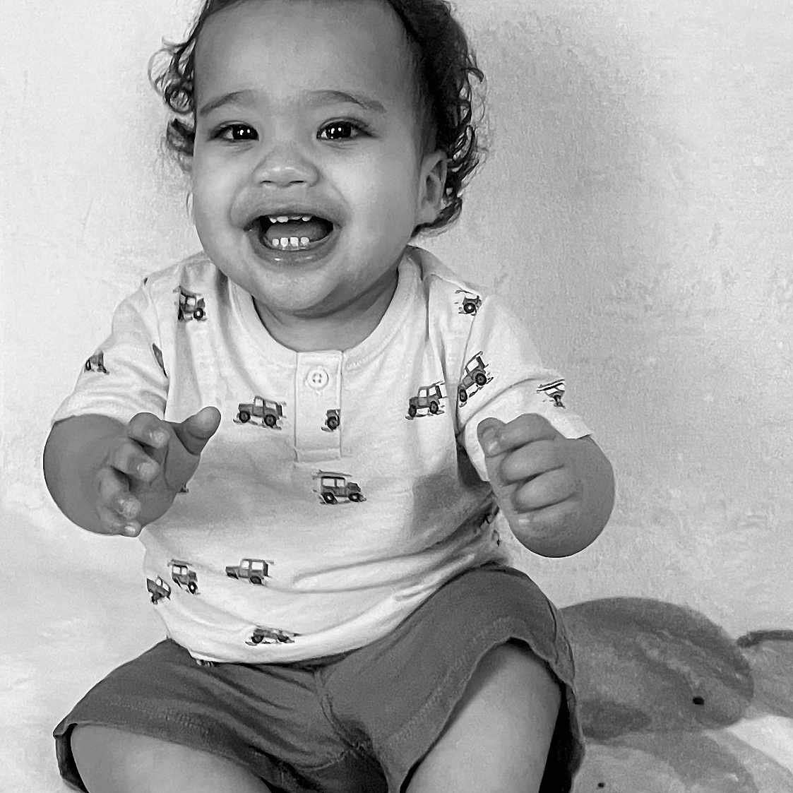 Jairo is registered to the contest to win money with this photo: baby, toddler, smiling, curly_hair, sitting, blanket, pattern, shorts, shirt, hand, feet, face, child, indoors, portrait, black_and_white, happy, cute, clothing, expression