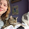Penelope is registered to the contest to win money with this photo: person, smiling, cat, tabby_cat, white_and_gray_cat, indoor, blanket, bed, purple_wall, painting, stitch_art, curtain, cozy, feline, pet, happy, young_adult, casual_clothing, portrait, relaxed