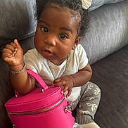 Anaria is registered to the contest to win money with this photo: toddler, child, curly_hair, white_bows, pink_purse, gray_couch, gray_pants, white_shirt, bracelet, necklace, sitting, indoor, cute, fashion, accessories, small_bag, person, cozy, portrait, curious