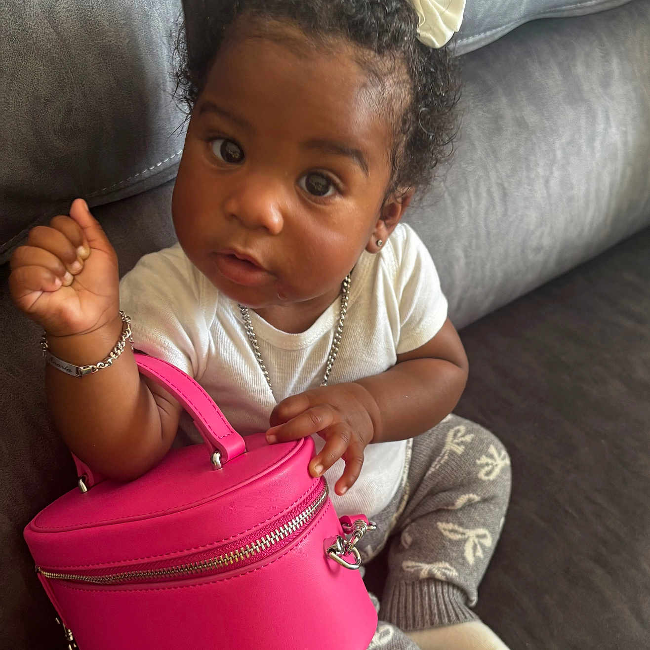 Anaria is registered to the contest to win money with this photo: accessories, bracelet, child, cozy, curious, curly_hair, cute, fashion, gray_couch, gray_pants, indoor, necklace, person, pink_purse, portrait, sitting, small_bag, toddler, white_bows, white_shirt