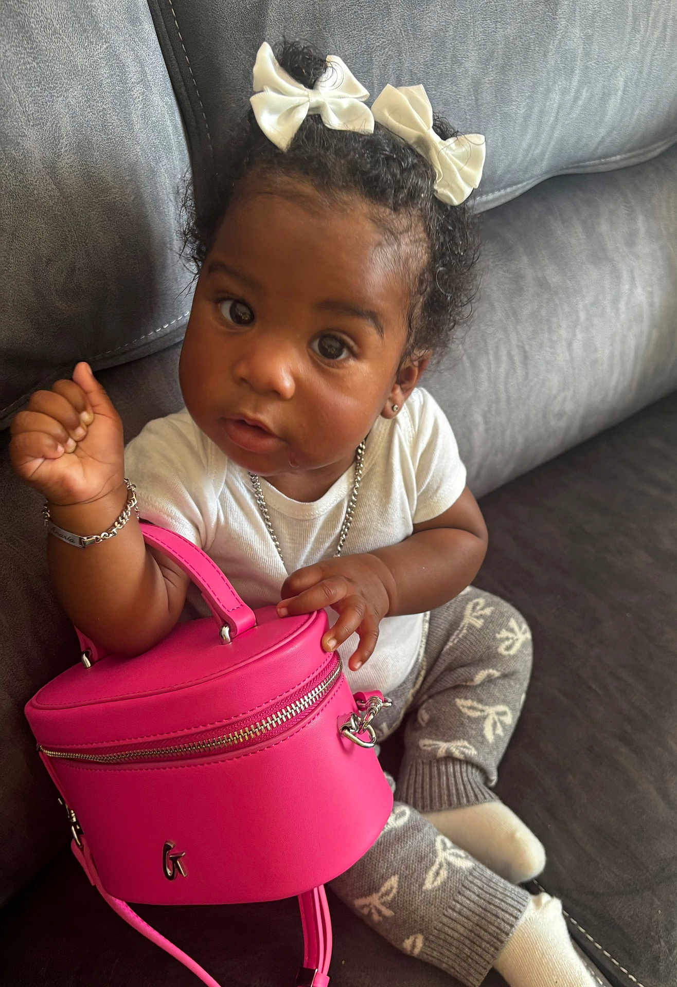 Anaria is registered to the contest to win money with this photo: toddler, child, curly_hair, white_bows, pink_purse, gray_couch, gray_pants, white_shirt, bracelet, necklace, sitting, indoor, cute, fashion, accessories, small_bag, person, cozy, portrait, curious