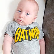 Logan participe au concours pour gagner de l'argent avec cette photo : baby, batman, blue_eyes, cartoon_character, casual_clothing, closeup, cute, gray_clothing, indoor, infant, lying_down, pants, person, portrait, relaxed, small_hand, soft_texture, tshirt, white_cushion, young_child