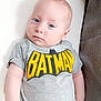 baby, batman, blue_eyes, cartoon_character, casual_clothing, closeup, cute, gray_clothing, indoor, infant, lying_down, pants, person, portrait, relaxed, small_hand, soft_texture, tshirt, white_cushion, young_child