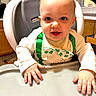 baby, child, clothing, curtains, eyes, face, green, hands, high_chair, indoor, infant, kitchen, shirt, sitting, smile, table, tray, window, wooden_cabinet, young_child