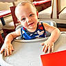 blue_eyes, chair, child, cute, daylight, face, furniture, hands, happy, high_chair, indoor, person, restaurant, skin, smiling, table, tank_top, toddler, window, wood_floor