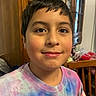 child, smile, face, indoor, wooden_furniture, cabinet, shirt, tie_dye, person, portrait, young, casual_clothing, happy, home, brown_hair, eyes, childhood, dining_room, furniture, closeup