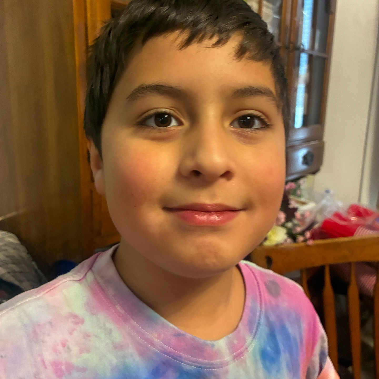 Mj is registered to the contest to win money with this photo: child, smile, face, indoor, wooden_furniture, cabinet, shirt, tie_dye, person, portrait, young, casual_clothing, happy, home, brown_hair, eyes, childhood, dining_room, furniture, closeup