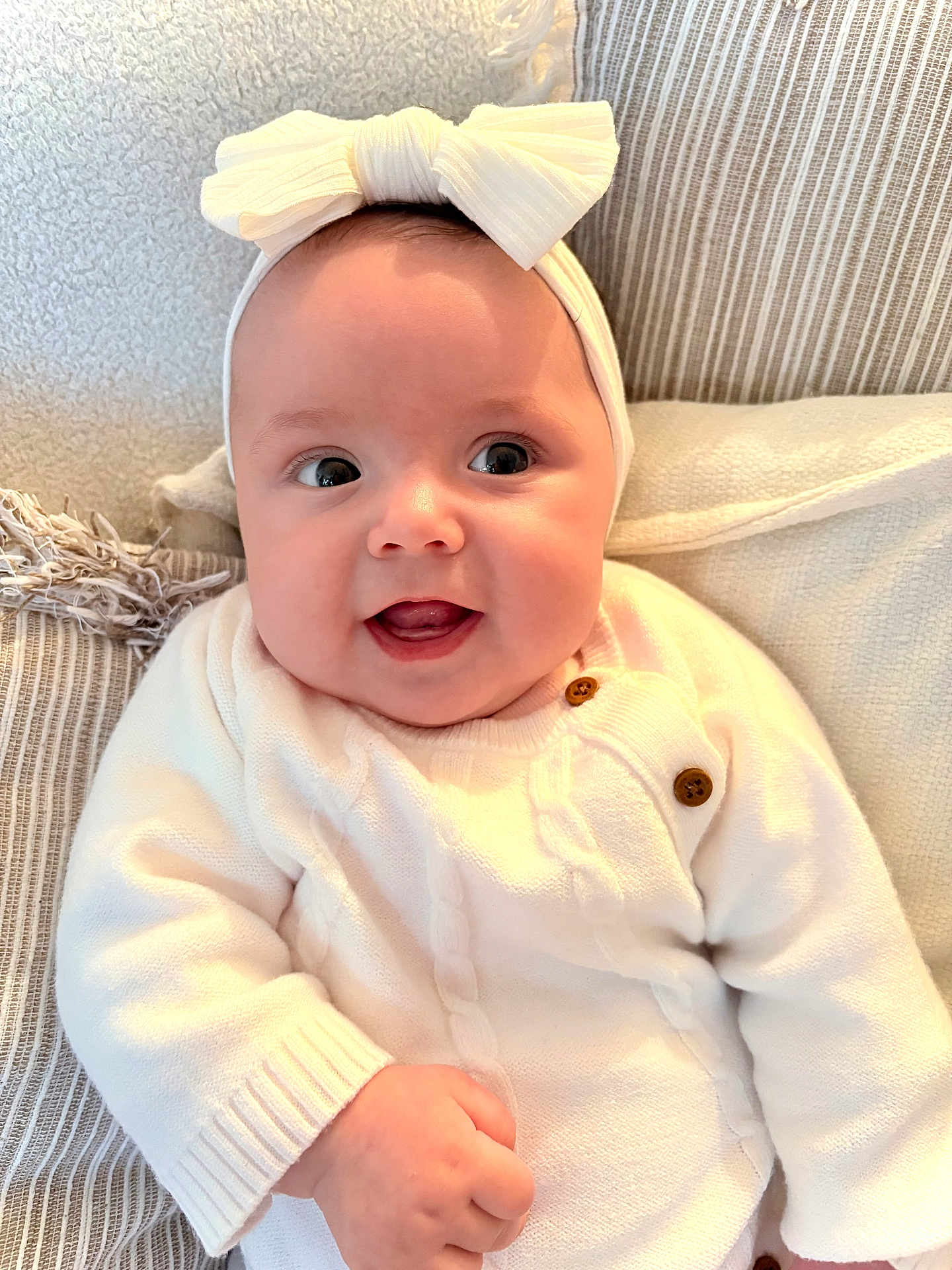 Dua is registered to the contest to win money with this photo: baby, child, smiling, headband, bow, white_sweater, couch, pillow, closeup, indoor, happy, infant, cute, portrait, soft_lighting, cozy, furniture, person, skin, hand