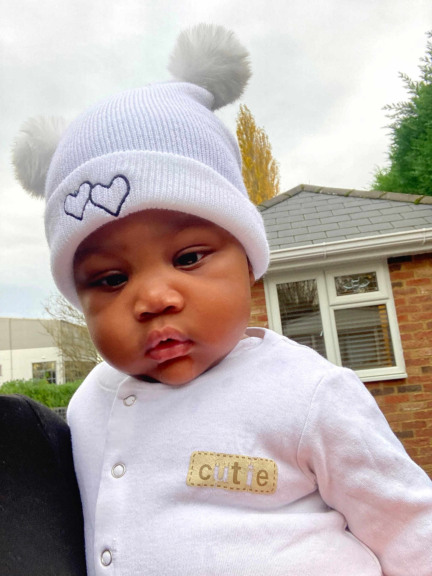 Miracle is registered to the contest to win money with this photo: baby, child, infant, white_hat, pom_poms, onesie, cutie_label, outdoor, suburban, brick_house, window, tree, autumn, chubby_cheeks, cute, portrait, face, person, clothing, headwear