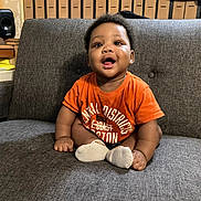 Miracle joined the competition — help win amazing prizes! baby, child, infant, smiling, orange_shirt, sitting, couch, indoor, furniture, happy, face, person, sock, wood_paneling, speaker, curly_hair, home, cute, young_child, casual