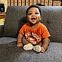 baby, child, infant, smiling, orange_shirt, sitting, couch, indoor, furniture, happy, face, person, sock, wood_paneling, speaker, curly_hair, home, cute, young_child, casual
