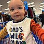 Liam is registered to the contest to win money with this photo: toddler, child, shopping_cart, shirt, clothing, indoor, store, retail, people, curious, bald, text_on_clothing, animal_graphics, long_sleeves, colorful, young_child, portrait, person, casual, happy