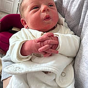Liam participe au concours pour gagner de l'argent avec cette photo : baby, infant, newborn, hands, clothing, white_outfit, person, indoor, cradled, face, expression, red_chair, cozy, soft_lighting, portrait, close_up, human, cute, child, warm