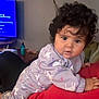baby, child, curly_hair, pajamas, unicorn_pattern, indoor, living_room, person, face, cute, curious, holding, red_clothing, furniture, television, table, bottle, hand, soft_lighting, cozy