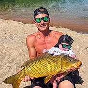 Levi is registered to the contest to win money with this photo: beach, lake, man, baby, fish, sunglasses, hat, sand, water, nature, outdoor, sunlight, reflection, summer, casual, fishing, child, adult, recreation, vacation