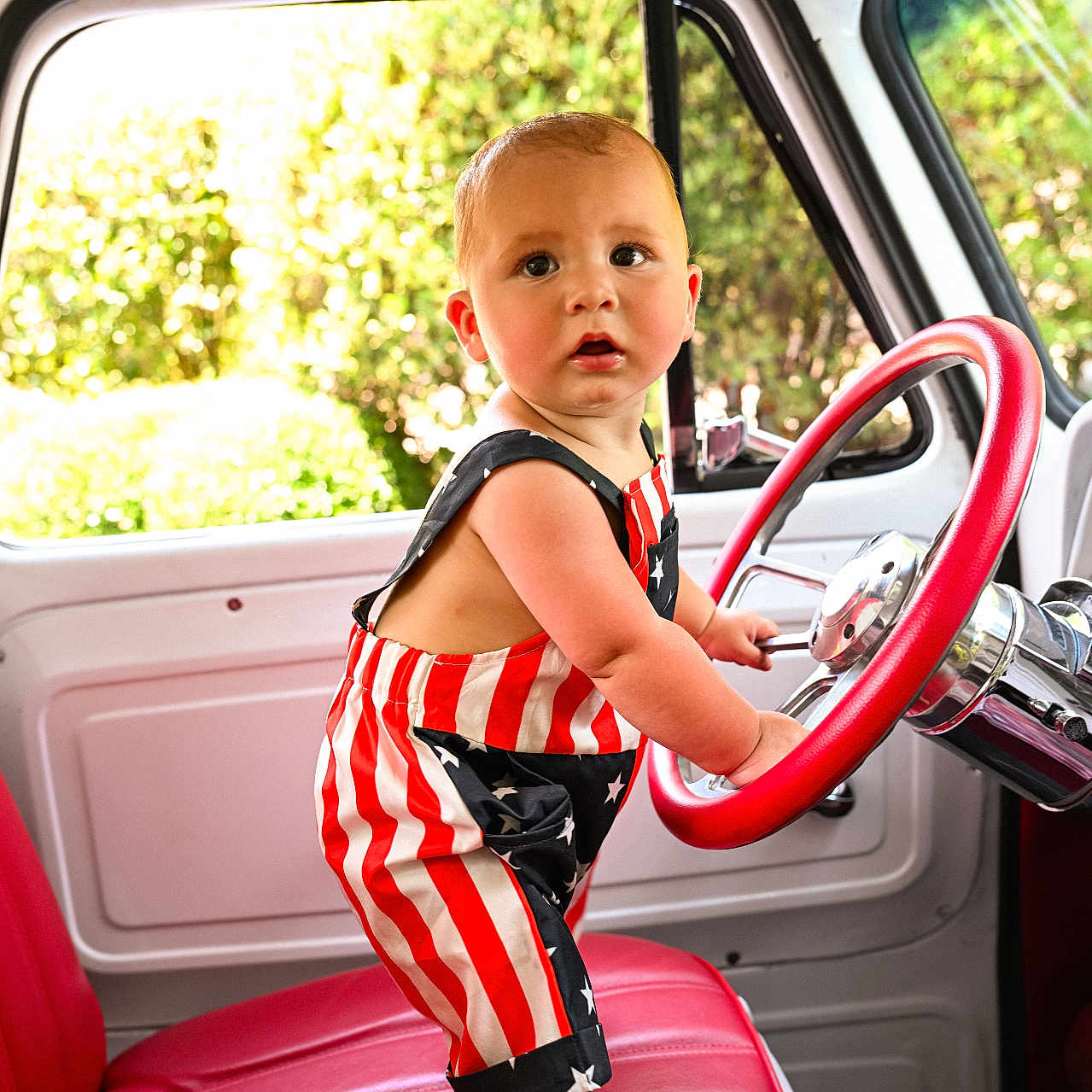 Levi joined the competition — help win amazing prizes! toddler, baby, child, car_interior, steering_wheel, red_seat, romper, american_flag_pattern, barefoot, window, daylight, greenery, vintage_car, curious_expression, indoor, portrait, person, seat, hand, face