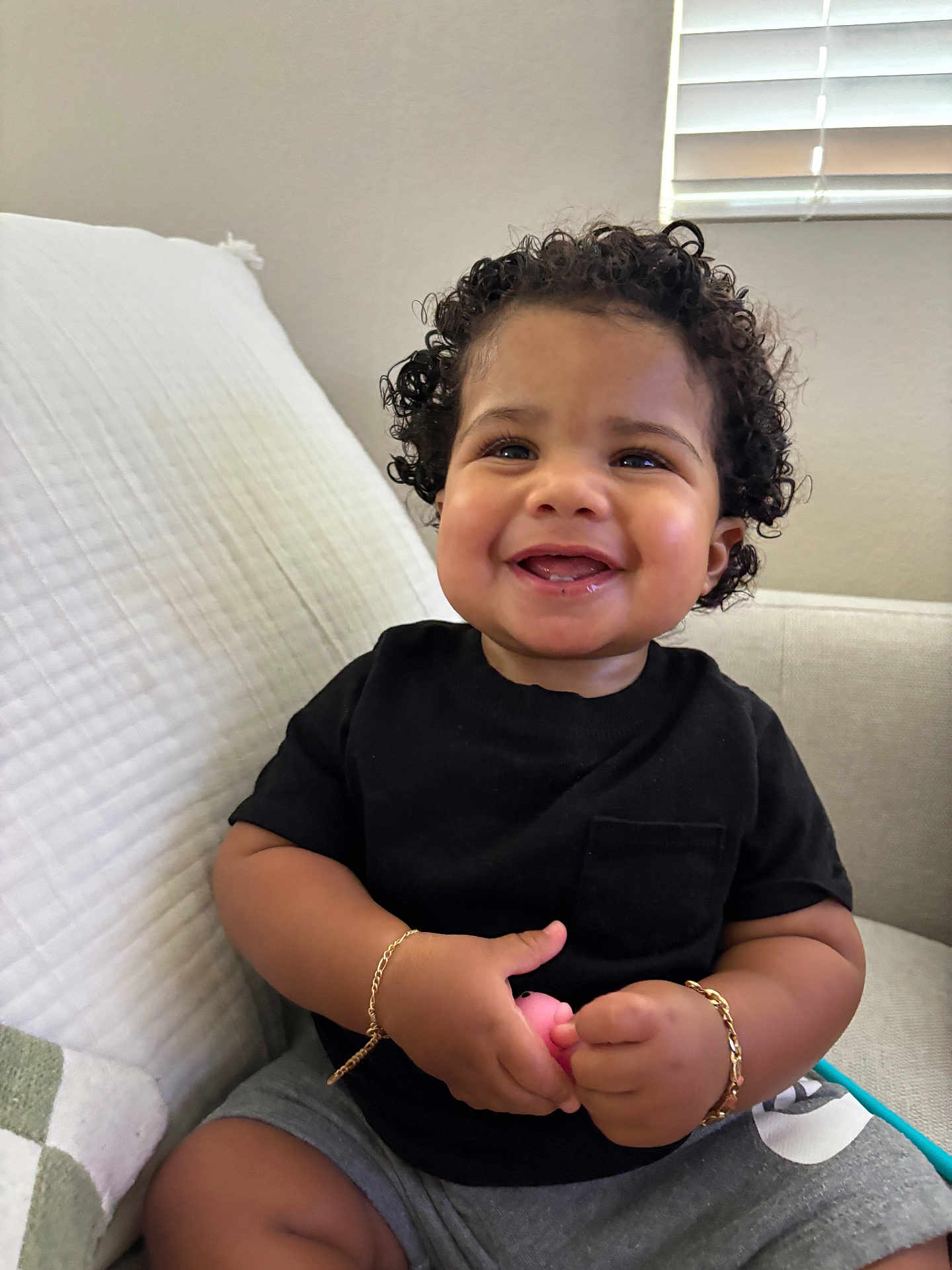 Alonis Saint Patrick joined the competition — help win amazing prizes! child, toddler, smiling, curly_hair, black_shirt, gray_shorts, bracelet, toy, couch, indoor, happy, cute, baby, person, sitting, portrait, home, window, light, skin