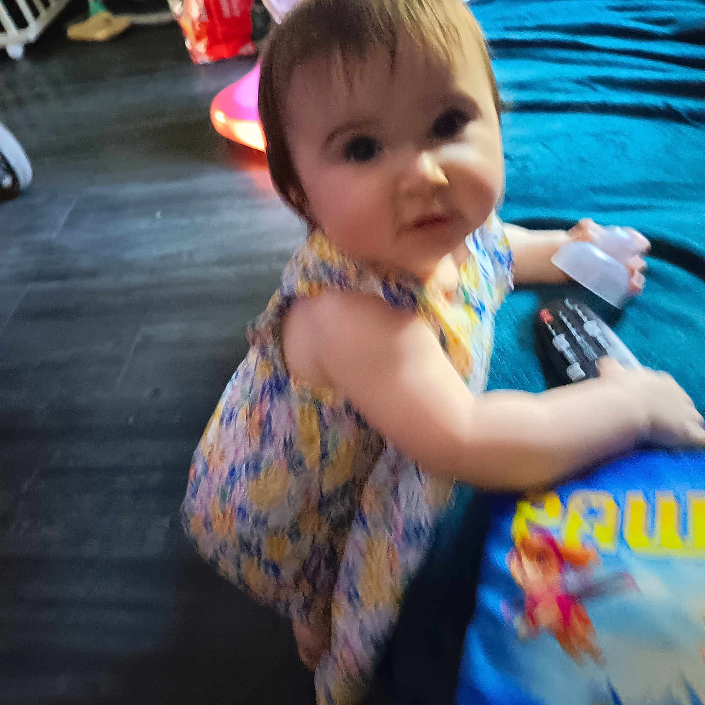 Mylèane a rejoint le concours — aidez-le/la à gagner de superbes lots ! blue_surface, bottle, cartoon_characters, child, curious, dress, face, floor, floral_pattern, furniture, hair, indoor, person, remote_control, room, sitting, skin, toddler, toy, young_child