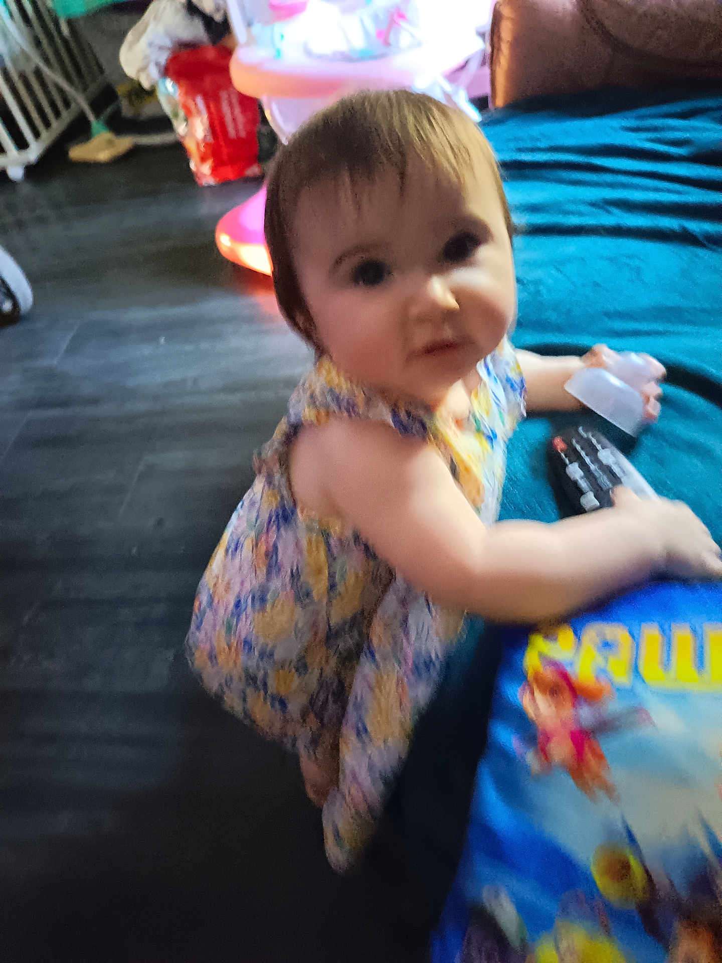 Mylèane a rejoint le concours — aidez-le/la à gagner de superbes lots ! toddler, child, indoor, dress, floral_pattern, bottle, blue_surface, cartoon_characters, face, curious, person, floor, toy, furniture, remote_control, hair, skin, sitting, young_child, room