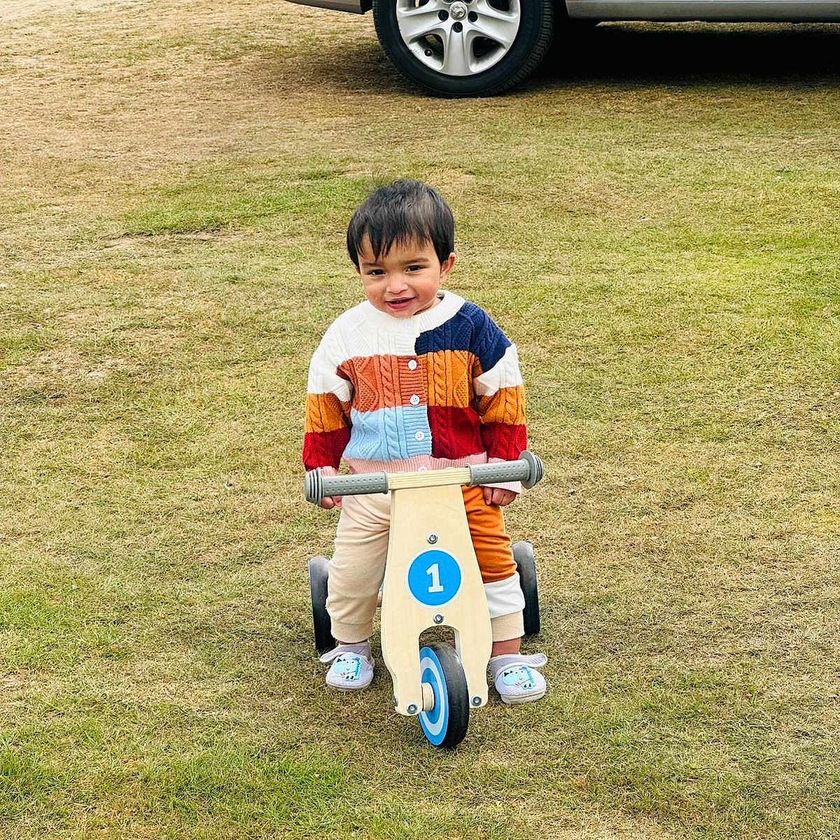 Johannes is registered to the contest to win money with this photo: balance_bike, car, casual_clothing, child, colorful_sweater, cute, daylight, footwear, grass, happy, nature, outdoor, park, play, smiling, toddler, toy, vehicle, wheel, young_child