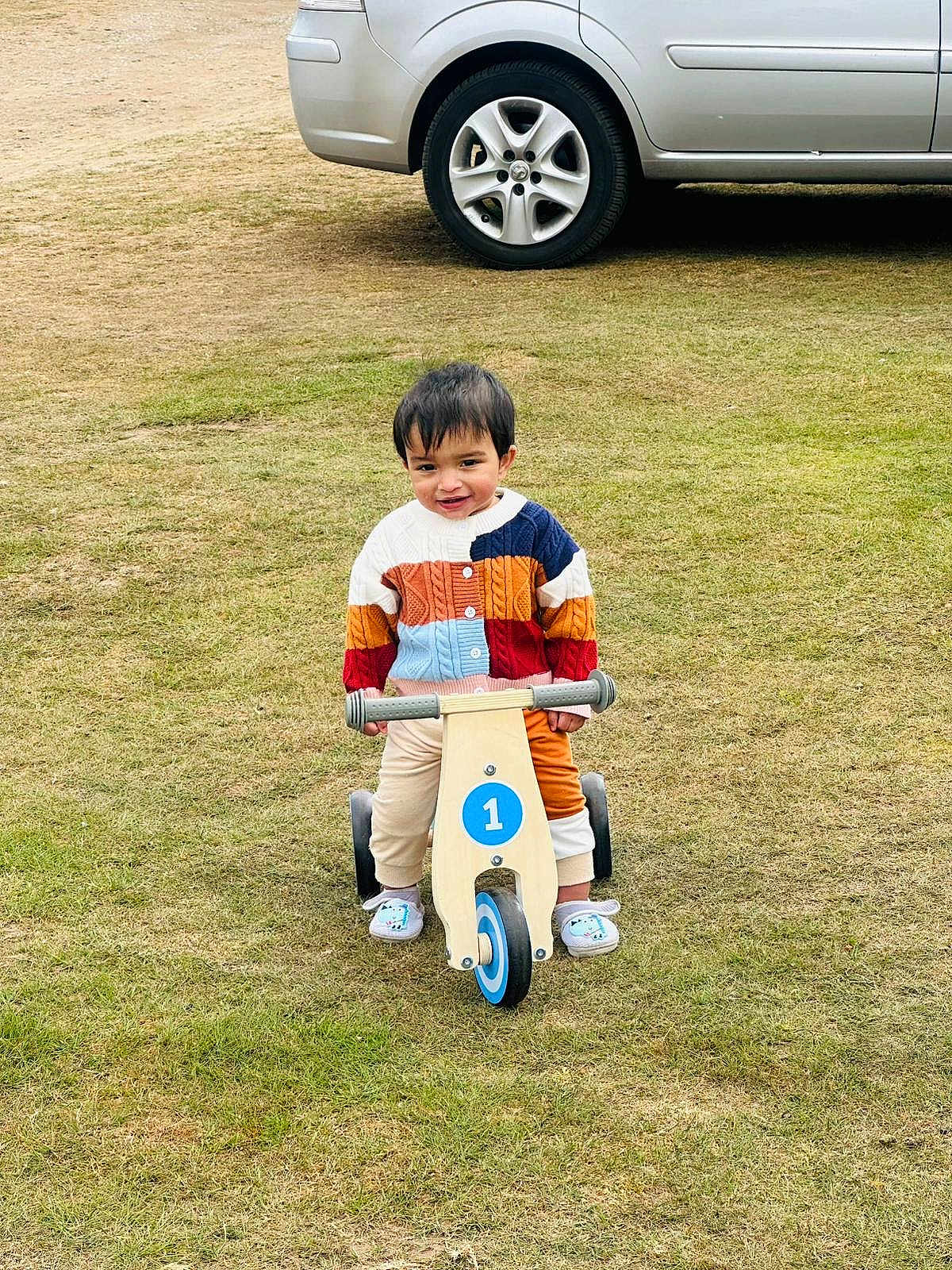 Johannes is registered to the contest to win money with this photo: toddler, child, balance_bike, colorful_sweater, grass, outdoor, toy, vehicle, car, wheel, footwear, happy, smiling, play, park, young_child, casual_clothing, daylight, nature, cute