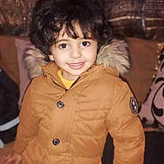 Ayden a rejoint le concours — aidez-le/la à gagner de superbes lots ! child, smiling, curly_hair, brown_jacket, fur_hood, indoor, cushions, curtains, person, portrait, happy, casual_clothing, young_child, warm_clothing, buttoned_jacket, seated, cute, face, eyes, hand