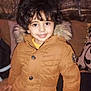 child, smiling, curly_hair, brown_jacket, fur_hood, indoor, cushions, curtains, person, portrait, happy, casual_clothing, young_child, warm_clothing, buttoned_jacket, seated, cute, face, eyes, hand