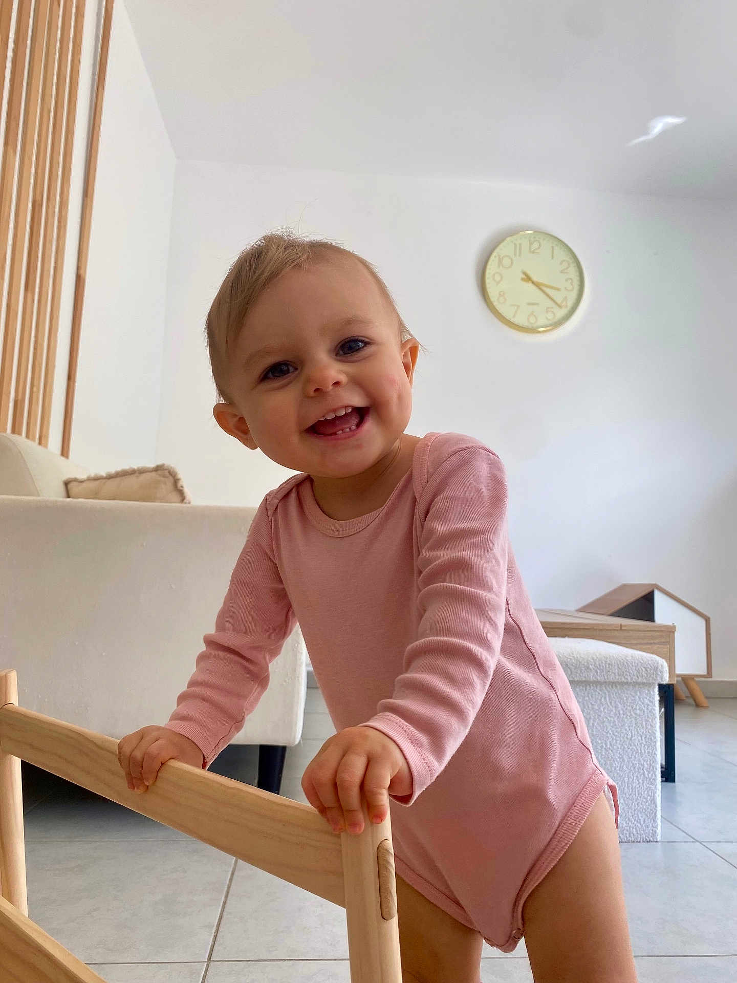 Estella participe au concours pour gagner de l'argent avec cette photo : toddler, child, smiling, pink_onesie, wooden_frame, indoor, modern_living_room, clock, floor_tiles, happy, dimples, baby, person, cute, furniture, natural_light, home, young_child, standing, playful