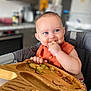 Aali joined the competition — help win amazing prizes! baby, child, high_chair, food, messy, blue_eyes, indoor, kitchen, bib, wooden_tray, hand, eating, smile, cute, infant, face, person, feeding, happy, mealtime