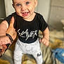 baby, child, smiling, blue_eyes, black_shirt, white_pants, hand, bed, indoor, person, happy, cute, standing, support, clothing, infant, face, portrait, human, background_blur