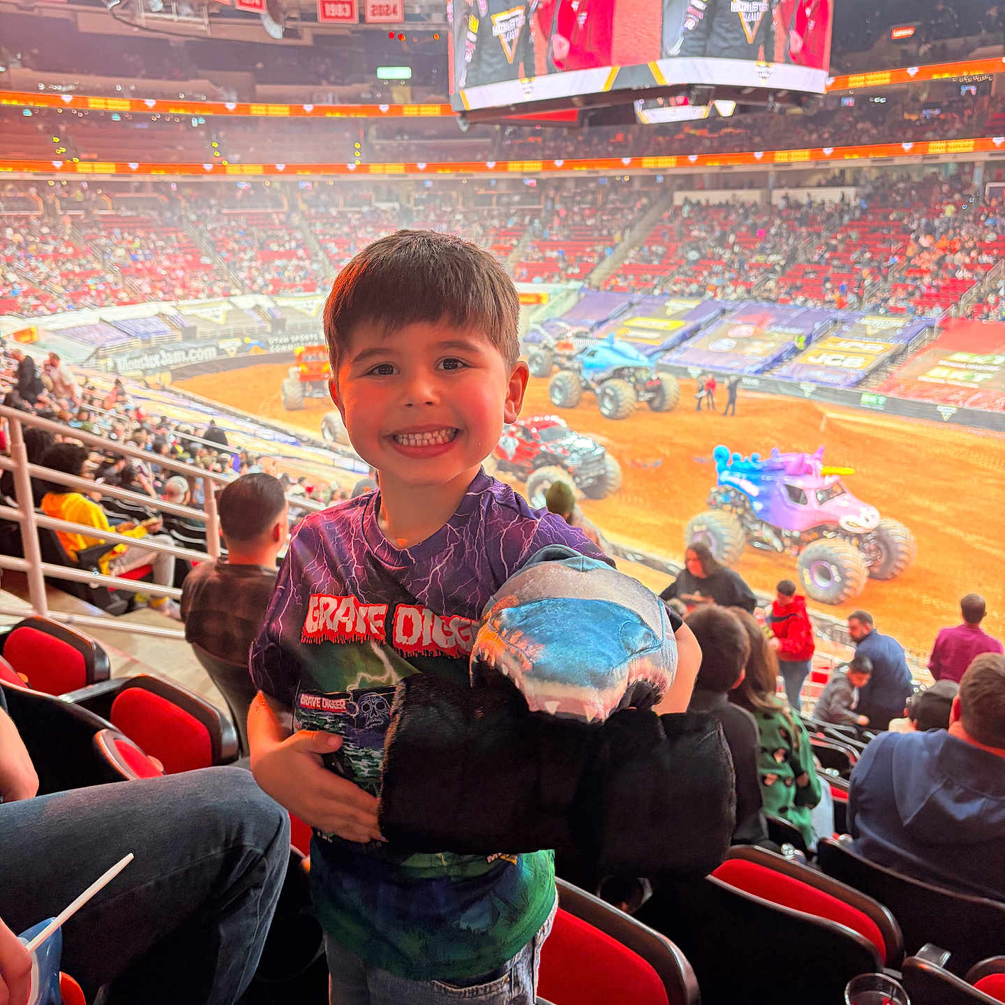 Conrad is registered to the contest to win money with this photo: arena, child, crowd, dirt_track, event, excited_face, family, jeans, monster_truck, plush_shark, railing, scoreboard, seats, smiling_child, spectators, stadium, stadium_lights, t_shirt, toy, vehicle