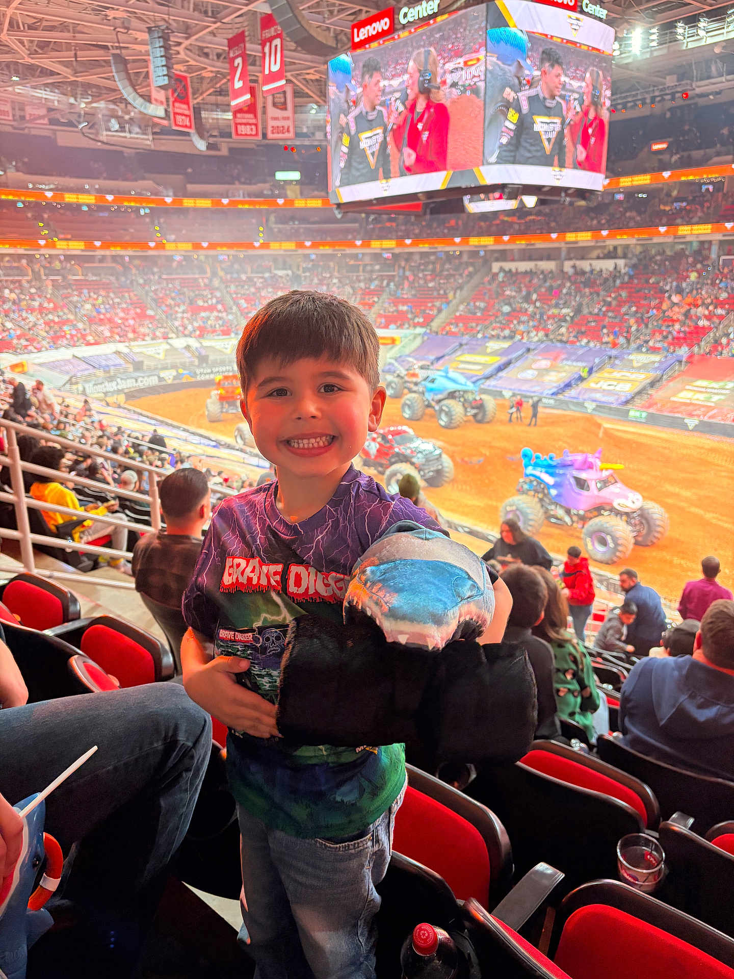 Conrad is registered to the contest to win money with this photo: child, smiling_child, plush_shark, monster_truck, stadium, arena, dirt_track, crowd, spectators, seats, scoreboard, stadium_lights, event, family, t_shirt, jeans, railing, toy, vehicle, excited_face